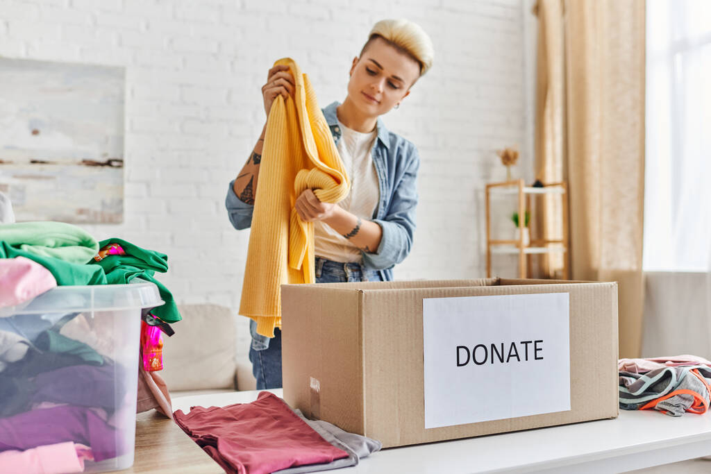 junge und stylische Frau in Freizeitkleidung mit gelbem Pullover neben Plastikbehälter mit Kleidungsstücken und Spendenbox im Wohnzimmer, nachhaltiges Leben und soziales Verantwortungskonzept - Foto, Bild