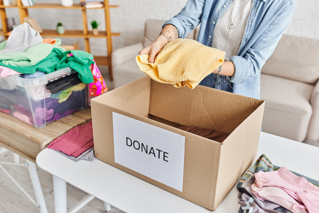 vista parcial de una mujer joven y tatuada sosteniendo un jersey amarillo encima de una caja de cartón con letras de donaciones cerca de un contenedor de plástico con ropa, vida sostenible y concepto de responsabilidad social - Foto, Imagen