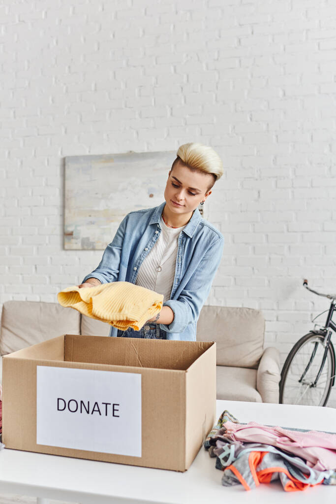 charita a dobrovolnictví, mladá tetovaná žena držící žlutý jumper v blízkosti šatní skříně a krabice s dárcovskými nápisy, koncepce udržitelného bydlení a společenské odpovědnosti - Fotografie, Obrázek