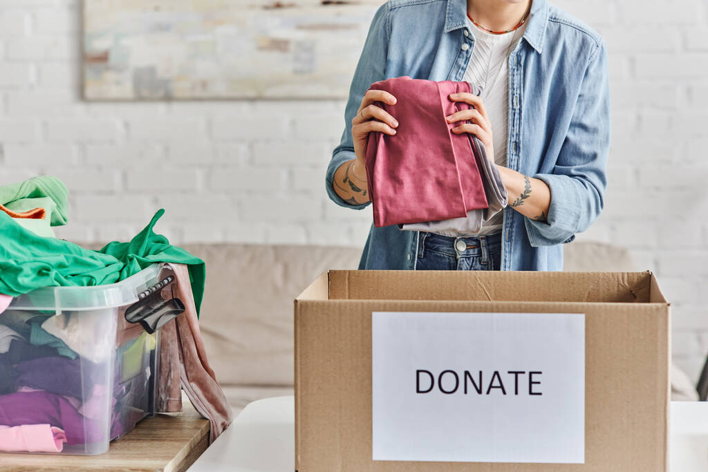 charité et bénévolat, vue recadrée d'une jeune femme tatouée tenant des vêtements de garde-robe près d'une boîte en carton avec lettrage de don, concept de vie durable et de responsabilité sociale - Photo, image