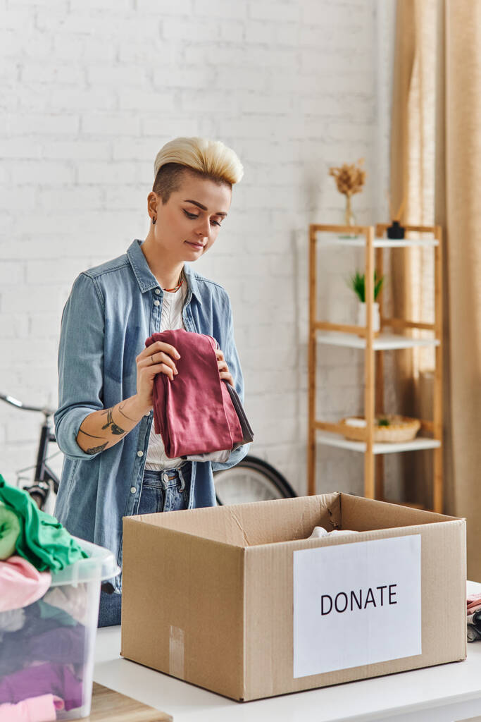 adományozás egy ügyért, tetovált nő divatos frizura gazdaság ruhák közelében kartondoboz adományozó betűkkel a modern nappaliban, fenntartható életmód és társadalmi felelősségvállalás koncepció - Fotó, kép