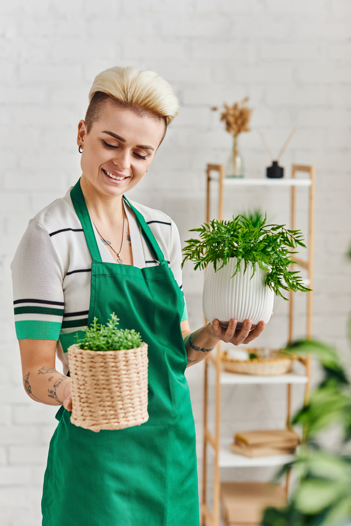 plant lover, eco-conscious lifestyle, pleased tattooed woman with radiant smile holding flowerpots with green plants in modern apartment, sustainable home decor and green living concept - Photo, Image