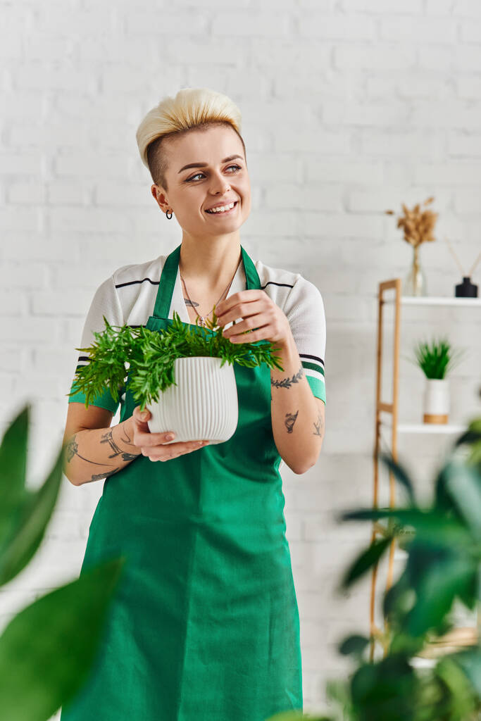 мечтательная и счастливая татуированная женщина в зеленом фартуке смотрит в сторону, стоя с горшочком в современной гостиной, размытый передний план, устойчивый декор дома и зеленая концепция жизни - Фото, изображение