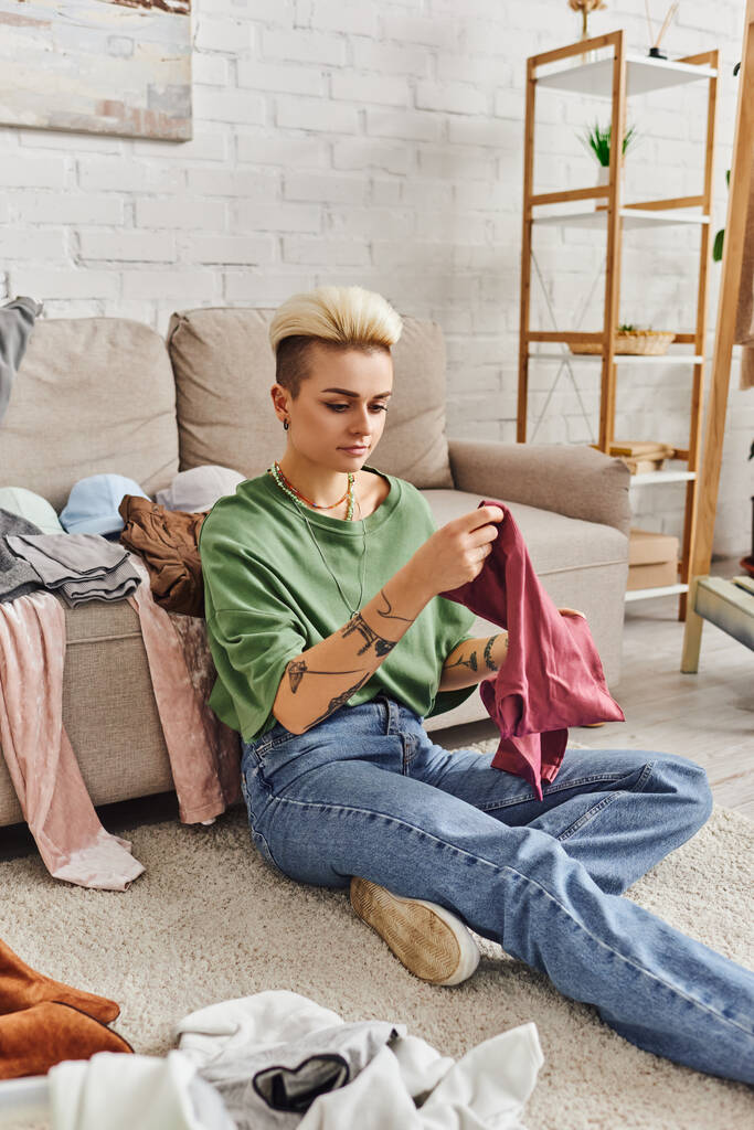ležérně stylová a tetovaná žena s módním účesem sedící na podlaze a třídějící použité předměty v blízkosti gauče v moderním obývacím pokoji, koncepce udržitelného bydlení a bdělého konzumerismu - Fotografie, Obrázek