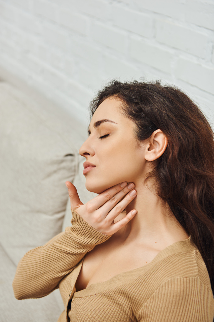 Young brunette woman in casual jumper massaging neck for lymphatic system support and relaxing on couch at home, self-care ritual and holistic wellness practices concept, tension relief - Photo, Image