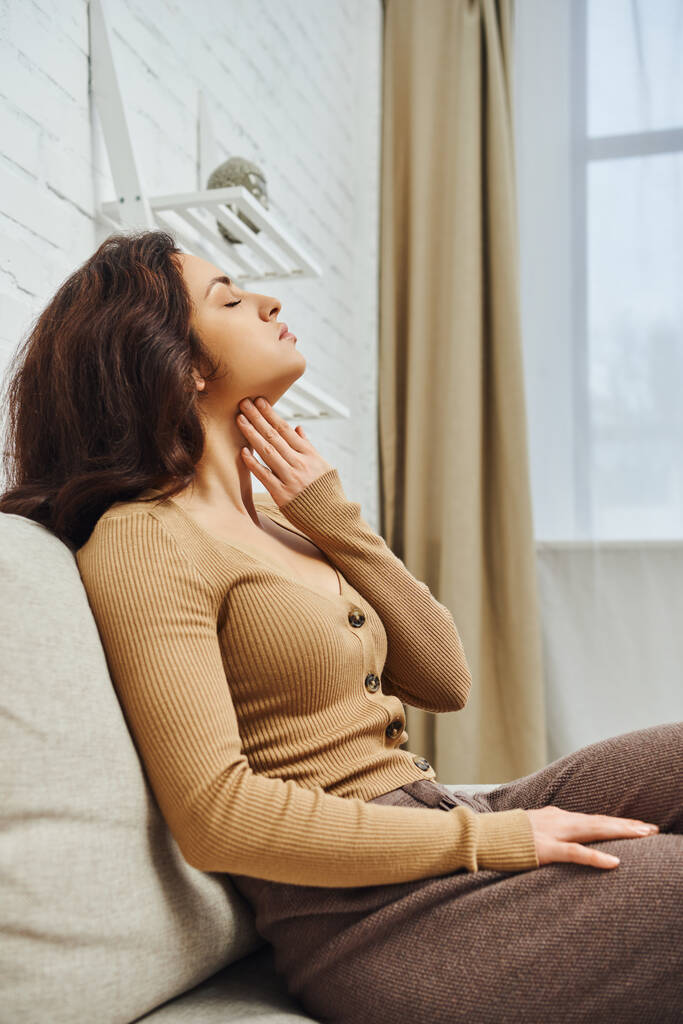 Side view of relaxed and young brunette woman in brown jumper touching neck during self-massage for lymphatic system on couch at home, self-care ritual and holistic wellness practices concept - Photo, Image