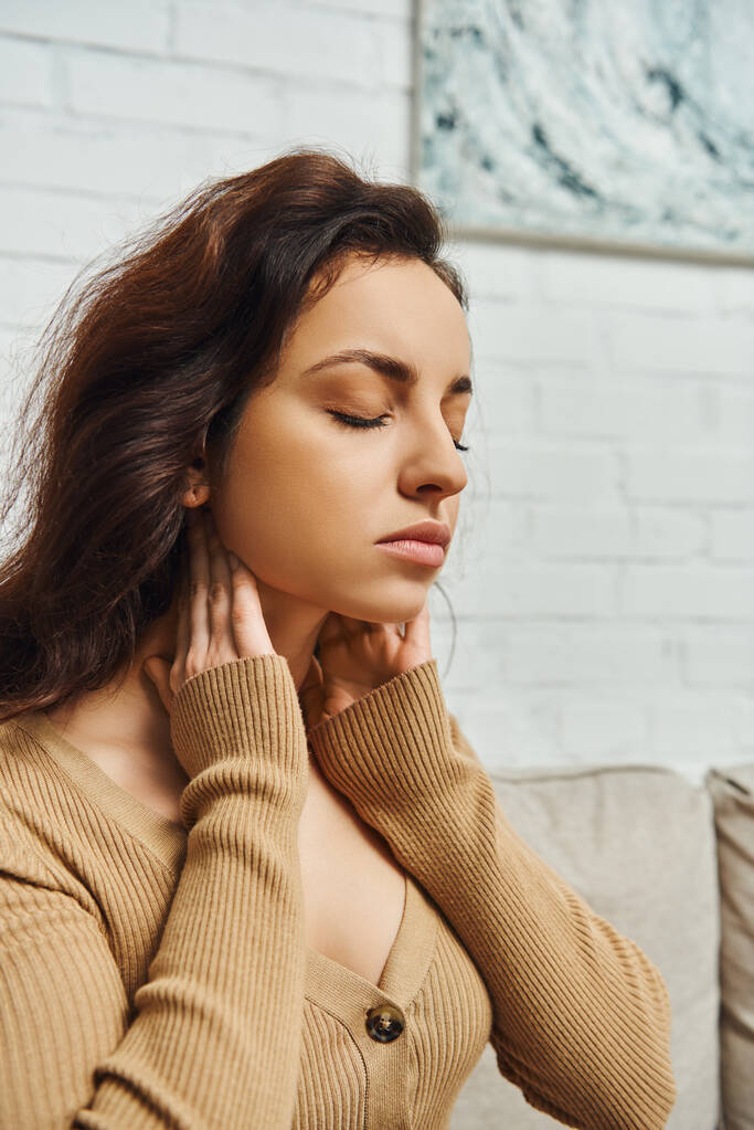 Portrait of young woman in casual brown jumper massaging lymphatic nodes and relaxing while sitting on blurred couch at home, self-care ritual and holistic wellness practices concept, tension relief - Photo, Image