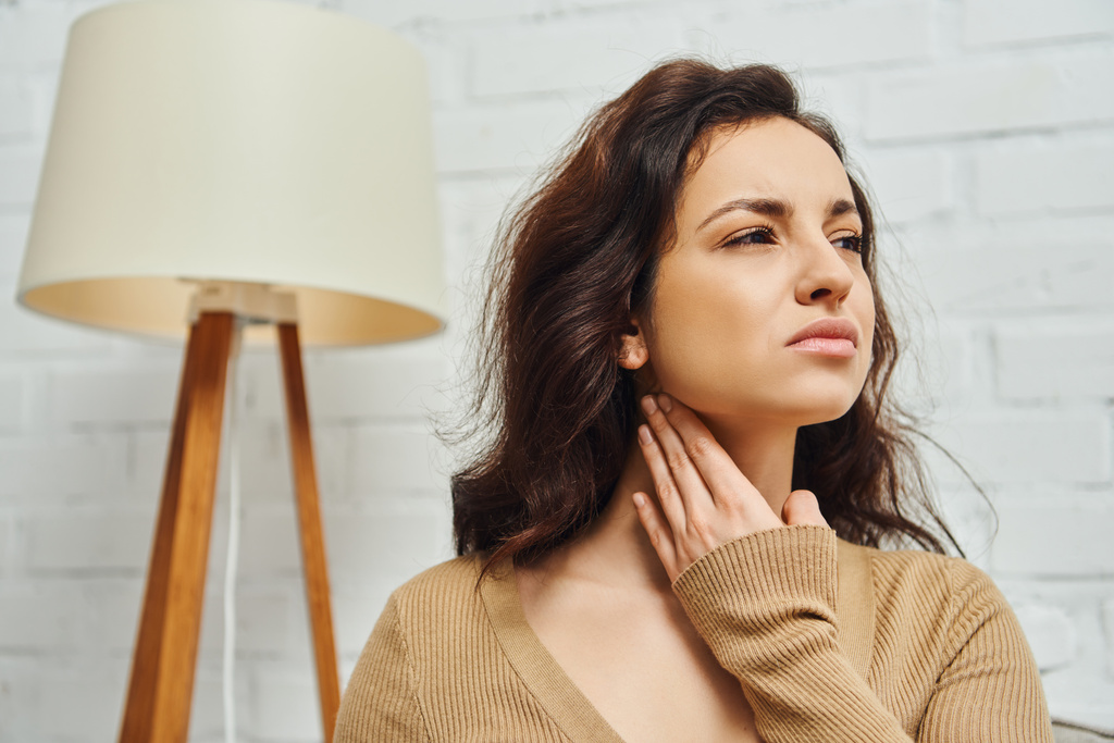 Displeased young brunette woman in brown jumper touching neck during lymphatic nodes self-massage at home, self-care ritual and holistic wellness practices concept, tension relief - Photo, Image