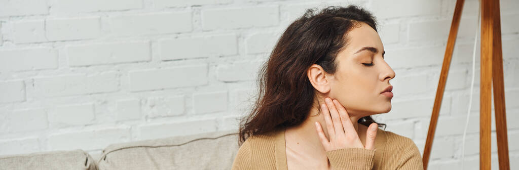 Side view of young brunette woman in casual brown jumper massaging lymphatic nodes on neck during self-massage on couch at home, self-care ritual and holistic healing concept, banner, tension relief - Photo, Image