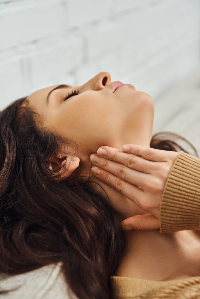 Mladá brunetka v hnědém svetru dotýkající se lymfatické uzliny na krku a relaxující při vlastní masáži doma na gauči, sebepéče rituální a holistická léčebná koncepce, uvolnění napětí - Fotografie, Obrázek