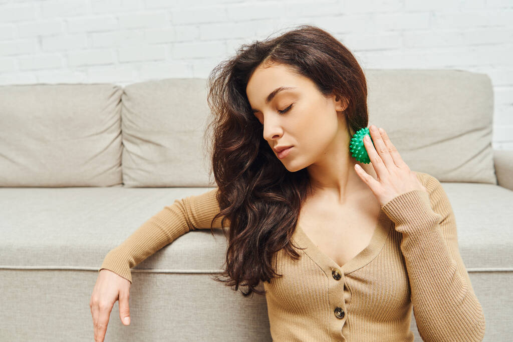 Young brunette woman in brown jumper massaging neck with manual massage ball while sitting near couch in living room, lymphatic system support and home-based massage, tension relief - Photo, Image
