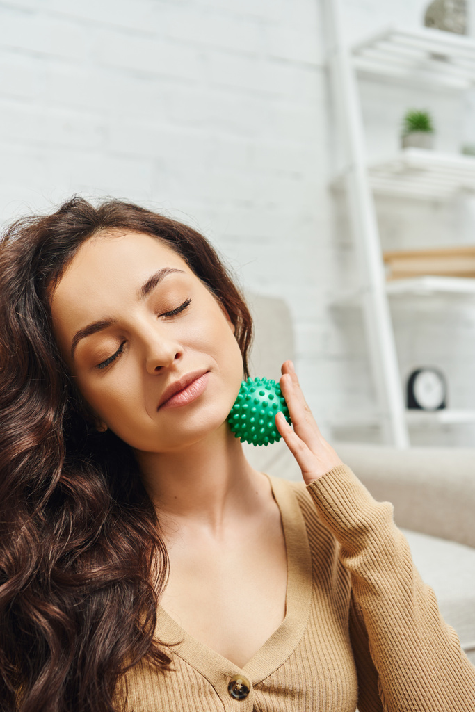 Porträt einer zufriedenen brünetten Frau mit geschlossenen Augen im braunen Pullover, die Lymphknoten mit manuellem Massageball im Wohnzimmer massiert, Lymphsystem unterstützt und zu Hause massiert - Foto, Bild