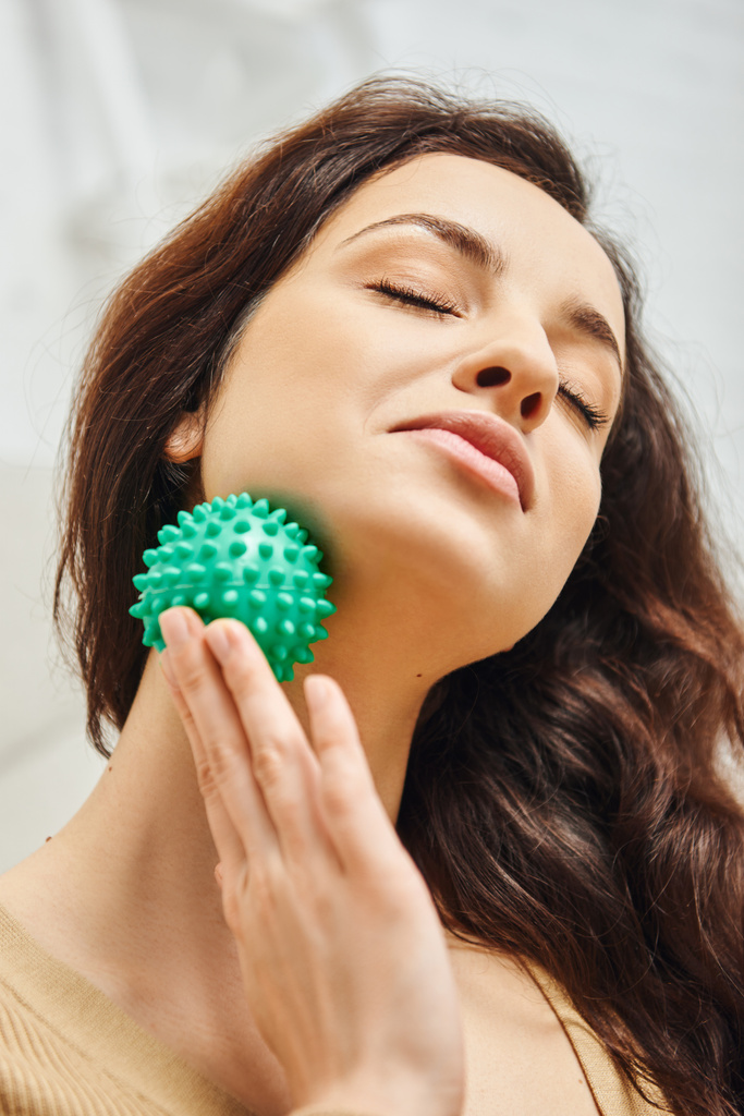 Portrait of pleased young brunette woman with closed eyes massaging neck with manual massage ball at home, lymphatic system support and home-based massage, balancing energy - Photo, Image