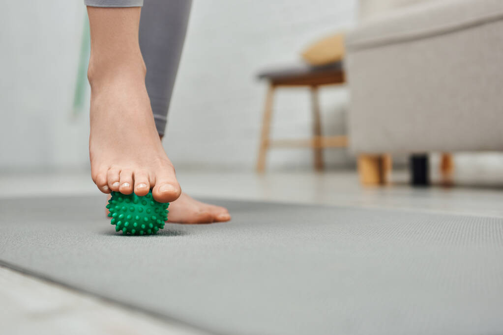 Vista recortada da mulher massageando o pé com bola de massagem manual e em pé no tapete de fitness em casa, relaxamento corporal e práticas de bem-estar holístico, equilibrando a energia - Foto, Imagem