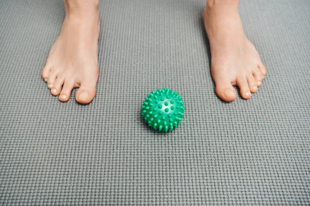 ruční masážní míč na fitness podložce u nohou ženy doma, relaxace těla a holistické wellness praktiky, vyvážení energetického konceptu, pohled shora, bosá noha  - Fotografie, Obrázek