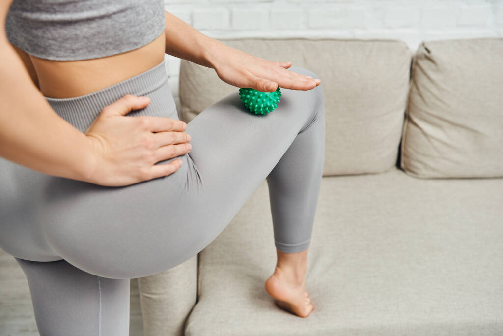 Cropped view of young woman in sportswear massaging leg with manual massage ball near couch in living room, holistic wellness practices and body relaxation concept, tension relief - Photo, Image