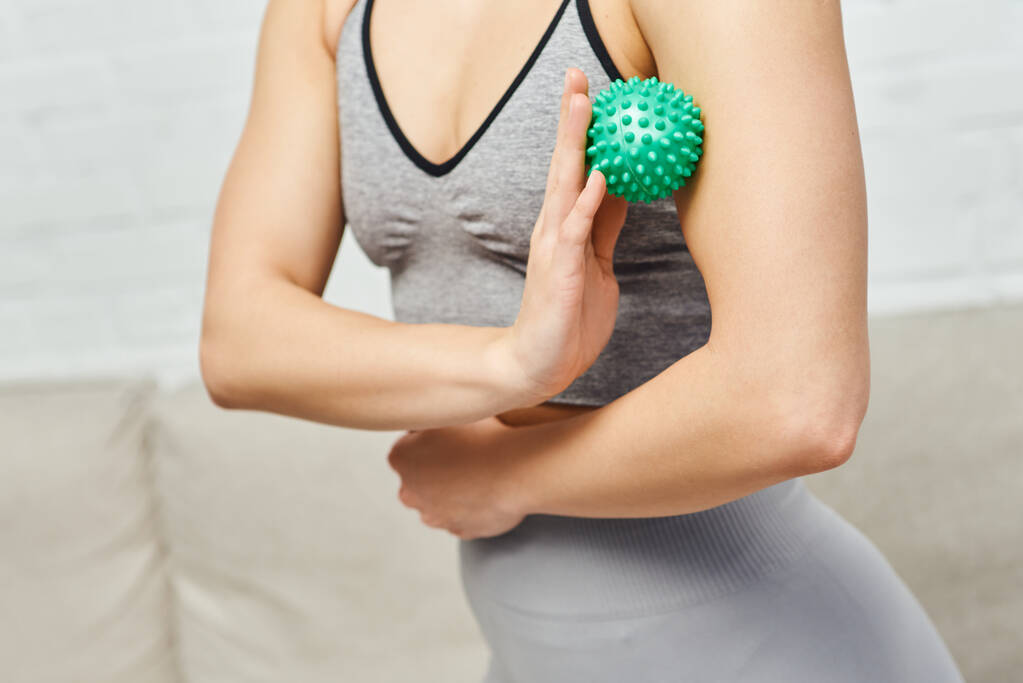 Обрезанный вид женщины в спортивной одежде массирующая мышца на руке с ручным массажным мячом в размытой гостиной, балансирующая энергия и целостная концепция исцеления, миофасциальное освобождение - Фото, изображение