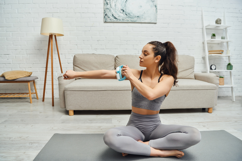 Young brunette woman in sportswear massaging arm with handled massager while sitting on fitness mat at home in living room, balancing energy and holistic healing concept, myofascial release - Photo, Image