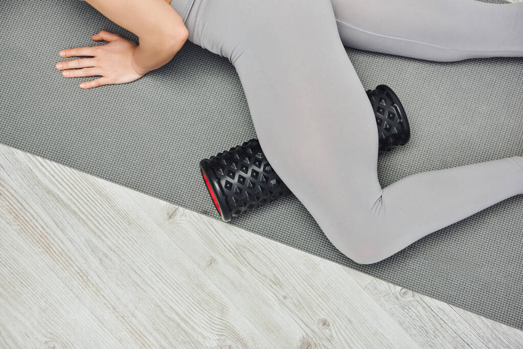 Обрезанный вид молодой женщины в активной одежде, массирующей ноги с помощью роллерного массажера и лежащей дома на коврике для фитнеса, акцент на концепции самообслуживания и благополучия, снятие напряжения - Фото, изображение