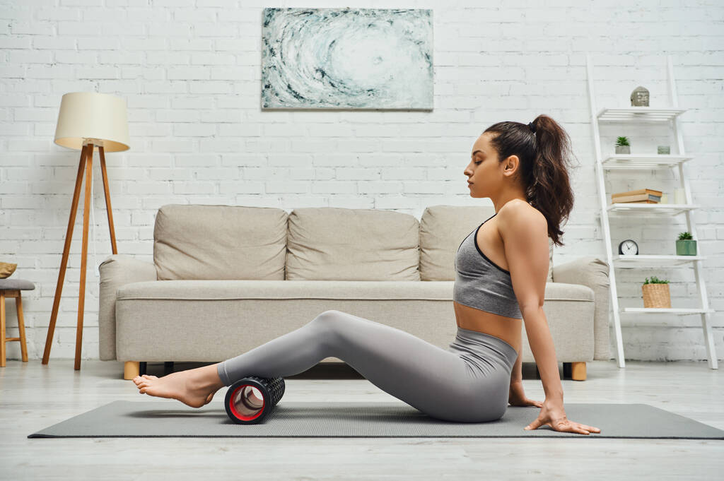 Side view of relaxed brunette woman in fitness clothes massaging legs with roller massager and sitting on mat in living room at home, maintaining healthy lymphatic system concept, myofascial release - Photo, Image