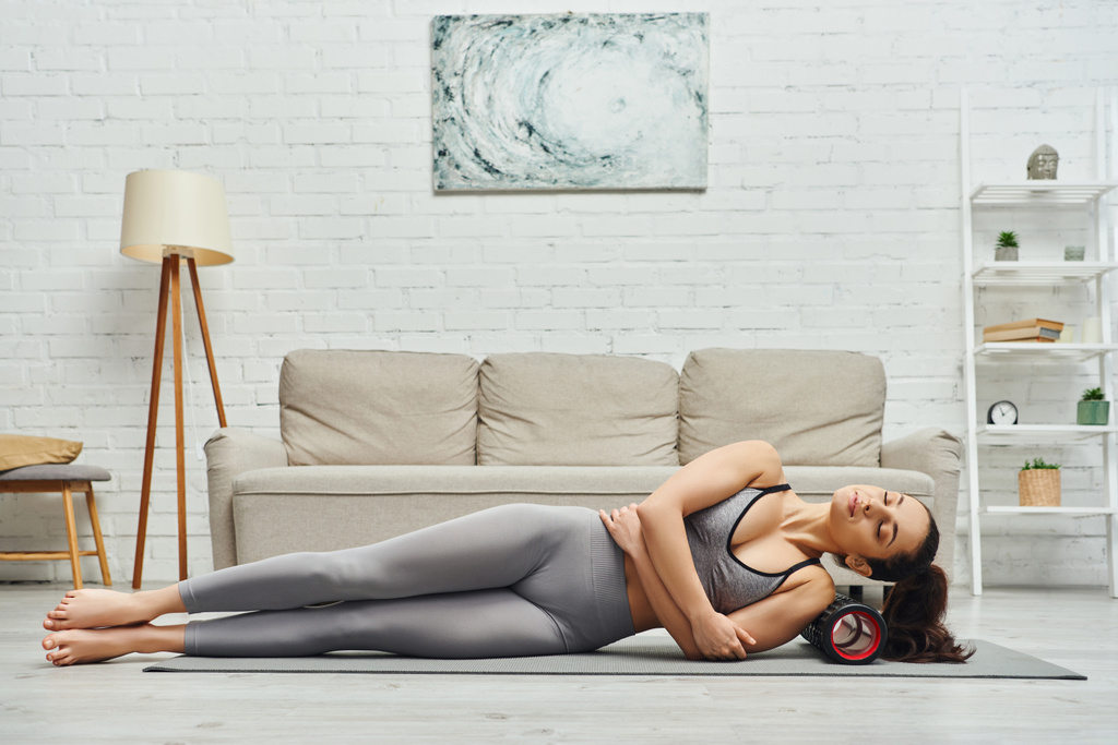Relaxed young woman in fitness clothes massaging shoulder with modern roller massager on mat in living room at home, sense of tranquility and promote relaxation concept, myofascial release - Photo, Image