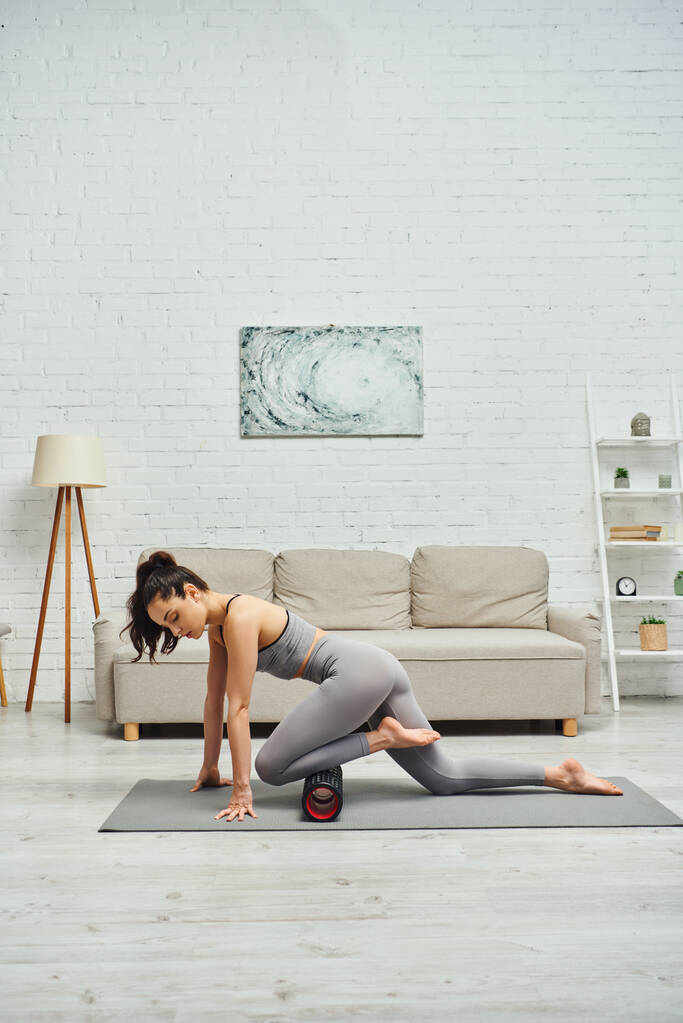 Junge brünette Frau in Aktivkleidung massiert Knie mit modernem Massageroller auf Fitnessmatte auf dem Boden im Wohnzimmer zu Hause, Gefühl der Ruhe und fördern Entspannungskonzept, myofasziale Befreiung - Foto, Bild