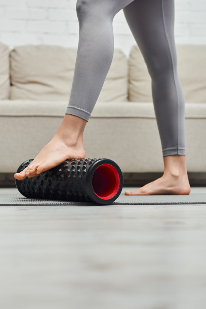 Widok na bosą kobietę w odzieży sportowej masujące stopy z nowoczesnym masażerem rolkowym na macie fitness w pobliżu kanapy w salonie, promowanie przepływu limfy i wellness w domu koncepcja, odciążenie napięcia - Zdjęcie, obraz
