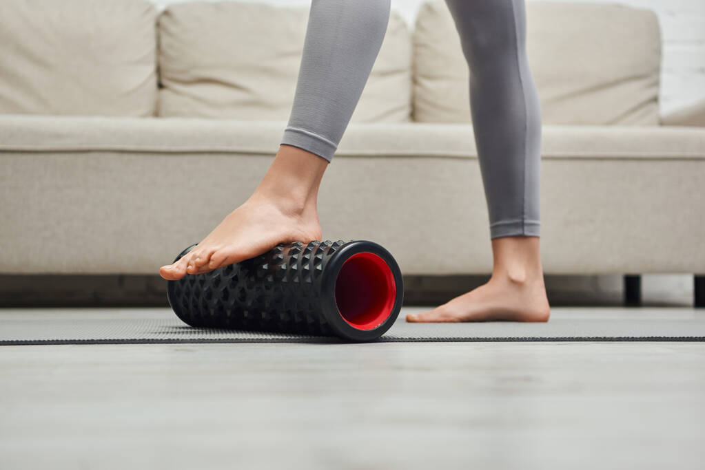 Cropped view of barefoot woman in sportswear massaging feet with roller massager and standing on fitness mat near couch at home, promoting lymph flow and wellness at home concept, tension relief - Photo, Image