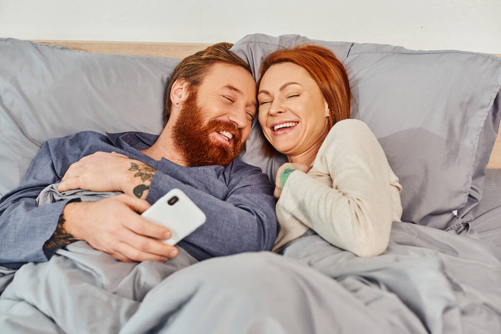 carefree, laughter, screen time, tattooed couple spending time without kids, day off, husband and wife, bearded man and redhead woman, smartphone users, cozy bedroom, home environment - Photo, Image