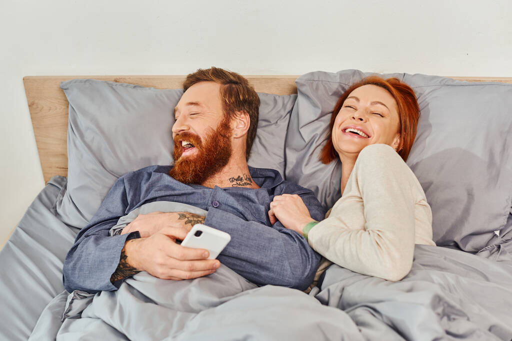 laughter, relaxation time, tattooed couple without kids, day off, husband and wife, bearded man and redhead woman, smartphone users, cozy bedroom, carefree, screen time  - Photo, Image