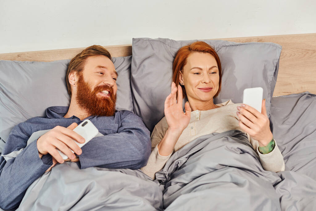 screen time, tattooed couple using smartphones, relaxing on weekends without kids, husband and wife, redhead woman having video call near bearded man, cozy bedroom, waving hand - Photo, Image