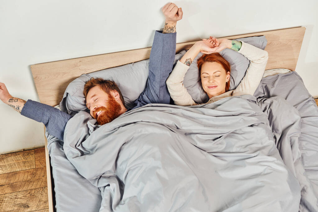 quiet house, parents alone at home, redhead husband and wife in cozy bedroom, bearded man and carefree woman relaxing on weekends, day off, wake up, tattooed, stretching, closed eyes, top view  - Photo, Image