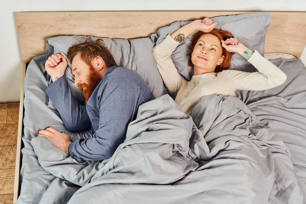 quiet house, parents alone at home, redhead husband and wife in cozy bedroom, bearded man sleeping near carefree woman relaxing on weekends, day off, wake up, tattooed, enjoy, top view  - Photo, Image
