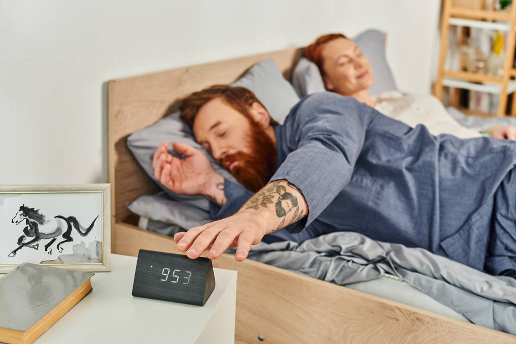 Uykulu sakallı adam elektronik alarma basıyor, çocuksuz bir gün, kızıl saçlı karı-koca, rahatlama zamanı, bulanık geçmiş, dövmeli, modern ev, rahat yatak odası.  - Fotoğraf, Görsel