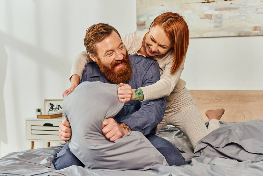 выходной день без детей, рыжая женщина обнимает бородатого мужа подушкой, счастливая пара во сне наслаждается временем вместе, татуированные люди, родители одни дома, качественное время вместе, игривый - Фото, изображение