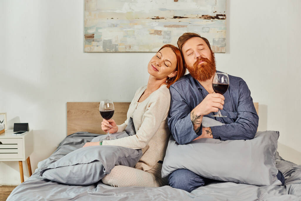 ρεπό χωρίς παιδιά, παντρεμένο ζευγάρι που κρατάει ποτήρια κόκκινο κρασί, κοκκινομάλλης σύζυγος και σύζυγος, απολαμβάνοντας χρόνο, ρεπό, σαββατοκύριακα μαζί, ευχαριστημένοι και με τατουάζ, γονείς μόνοι στο σπίτι  - Φωτογραφία, εικόνα