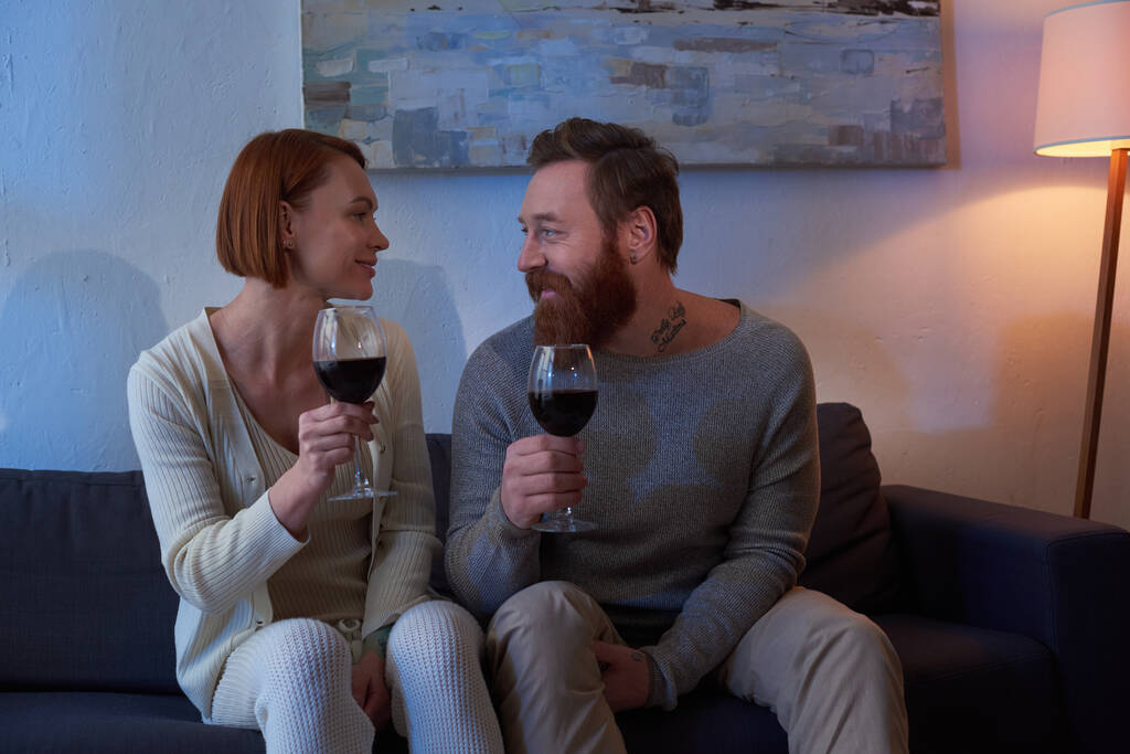 quality time, couple without kids enjoying time at home, tattooed couple relaxing on weekends, holding glasses of red wine, husband and wife, bearded man and redhead woman, cozy living room, day off  - Photo, Image