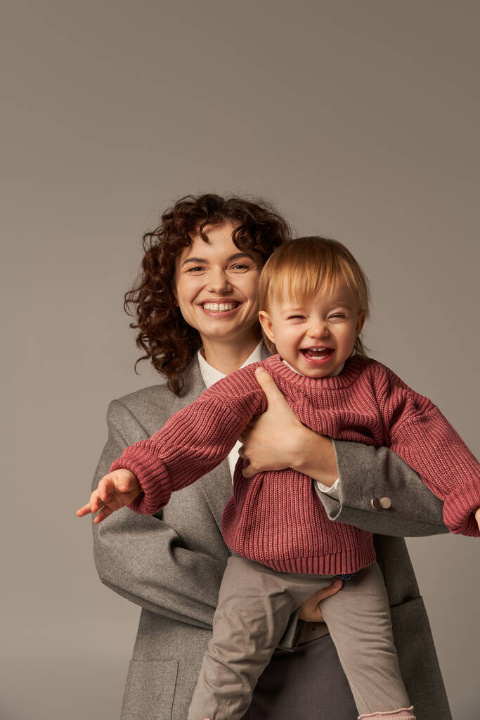 quality time, career and family, smiling woman in suit holding daughter on grey background, career and family, loving motherhood, lifestyle, multitasking, work life balance concept  - Photo, Image