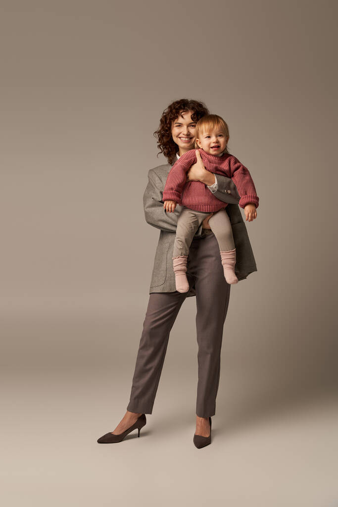 quality time, career and family, happy woman in suit holding child on grey background, growth and family, loving motherhood, lifestyle, multitasking, work life harmony concept  - Photo, Image