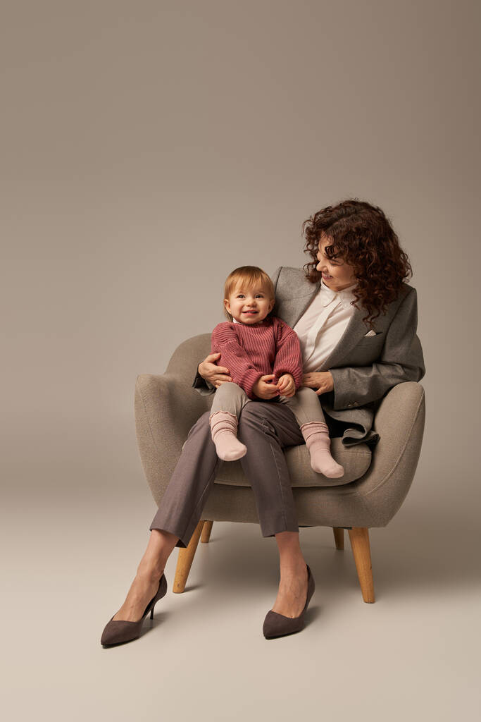 nowoczesny pracujący rodzic, równoważenie pracy i koncepcji życia, kręcone kobieta w garniturze siedzi w fotelu z małą córeczką, szare tło, szczęśliwa matka i dziecko, wielozadaniowość, jakość czasu  - Zdjęcie, obraz