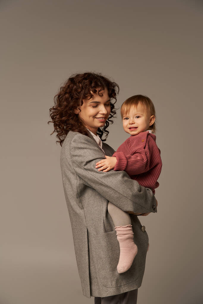 quality family time, parenting and career, cheerful businesswoman holding in arms her toddler daughter on grey background, work life harmony concept, working parent, loving motherhood - Photo, Image