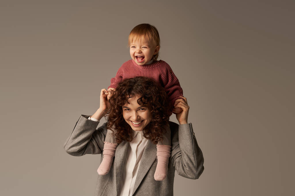 motherhood, excited kid on shoulders, work life balance, cheerful working mother, woman and happy daughter on grey background, work life harmony concept, loving motherhood, quality family time  - Photo, Image