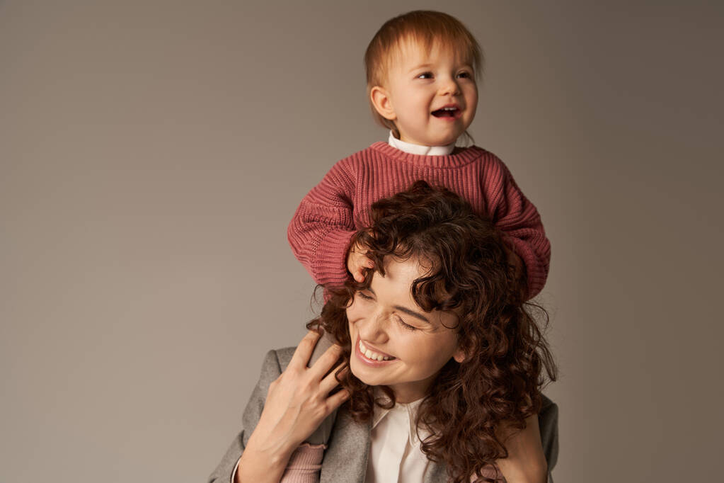 motherhood, kid on shoulders, work life balance, curly businesswoman and happy daughter on grey background, work life harmony concept, loving motherhood, quality family time  - Photo, Image