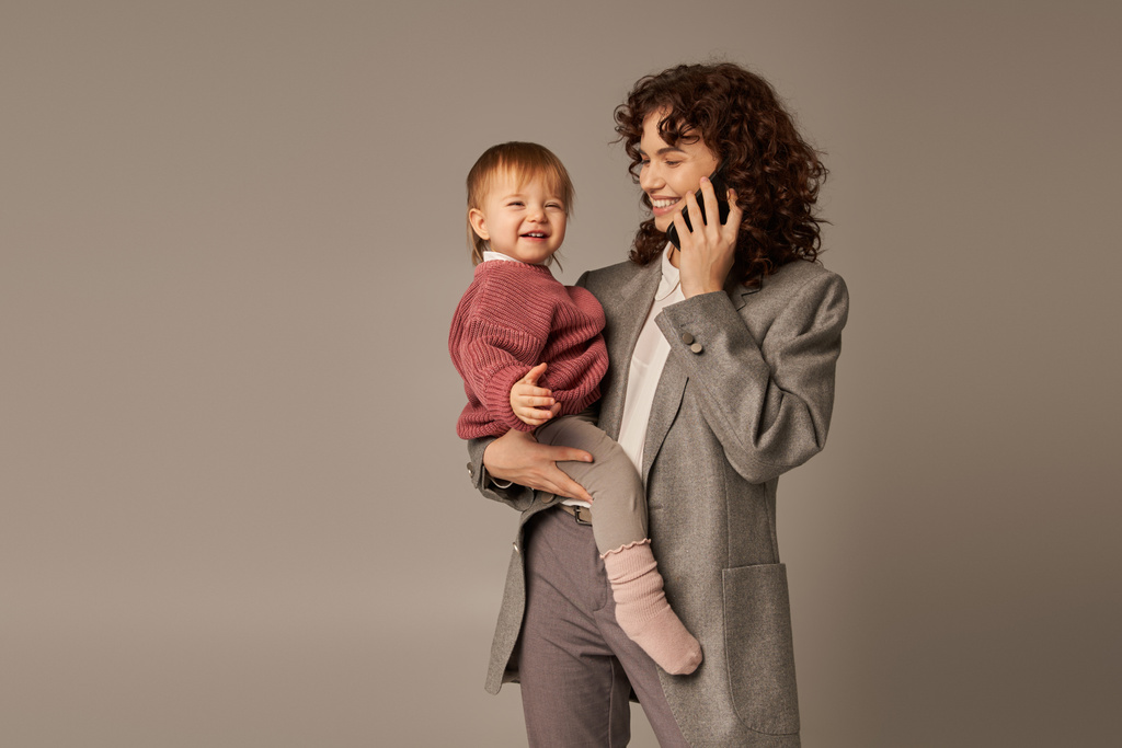 nowoczesne rodzicielstwo, budowanie udanej kariery, upodmiotowiona kobieta, wesoła matka rozmawiająca na smartfonie i trzymająca w ramionach córeczkę maluszka na szarym tle, macierzyństwo, businesswoman  - Zdjęcie, obraz