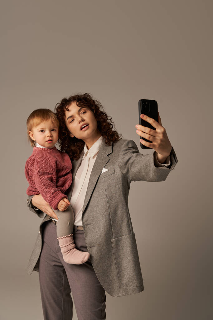 crianza moderna, construir una carrera exitosa, mujer empoderada, madre rizada tomando selfie en el teléfono inteligente y sosteniendo en brazos hija pequeña sobre fondo gris, maternidad  - Foto, Imagen