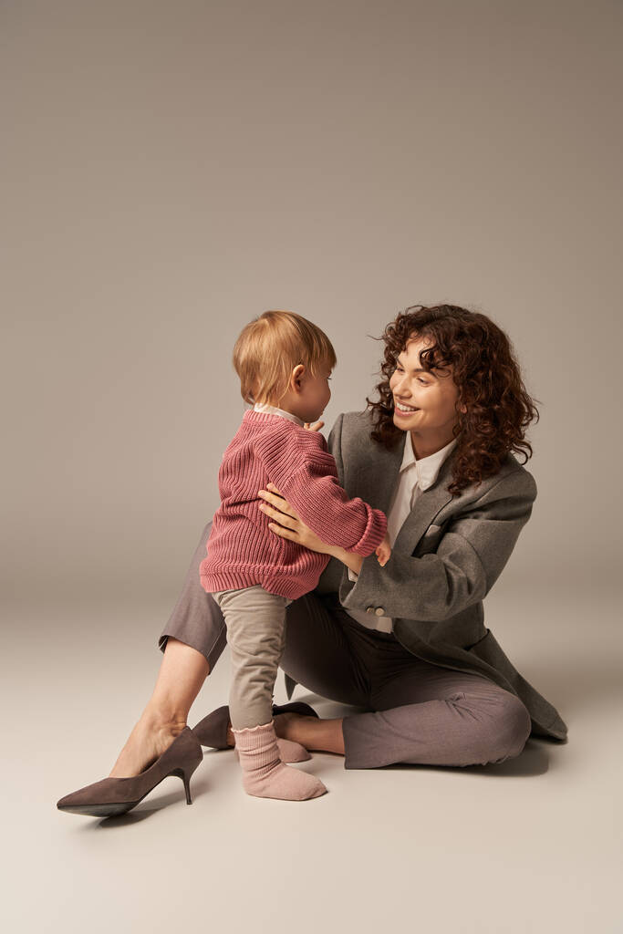 career and family, balancing between work and life, cheerful woman with curly hair hugging toddler daughter on grey background, quality time, modern parenting, motherhood, full length   - Photo, Image
