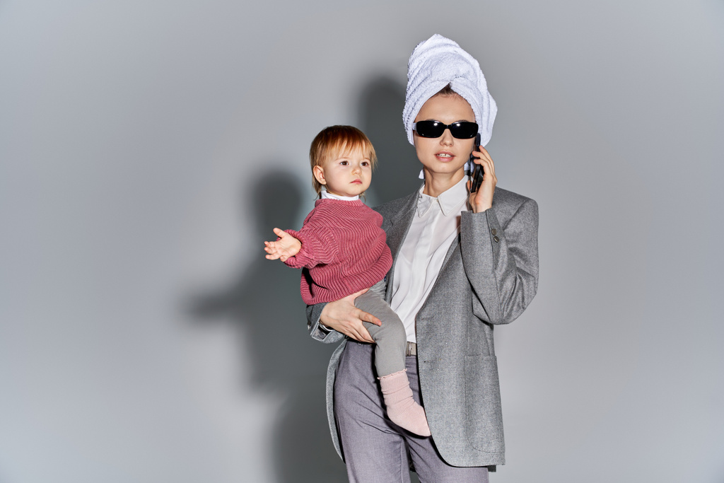 マルチタスク、腕の中で保持サングラスの女性幼児の娘と頭の上にタオルで立って、ライフスタイルのバランスをとる、灰色の背景にスマートフォンで話す正式な摩耗のビジネス女性  - 写真・画像