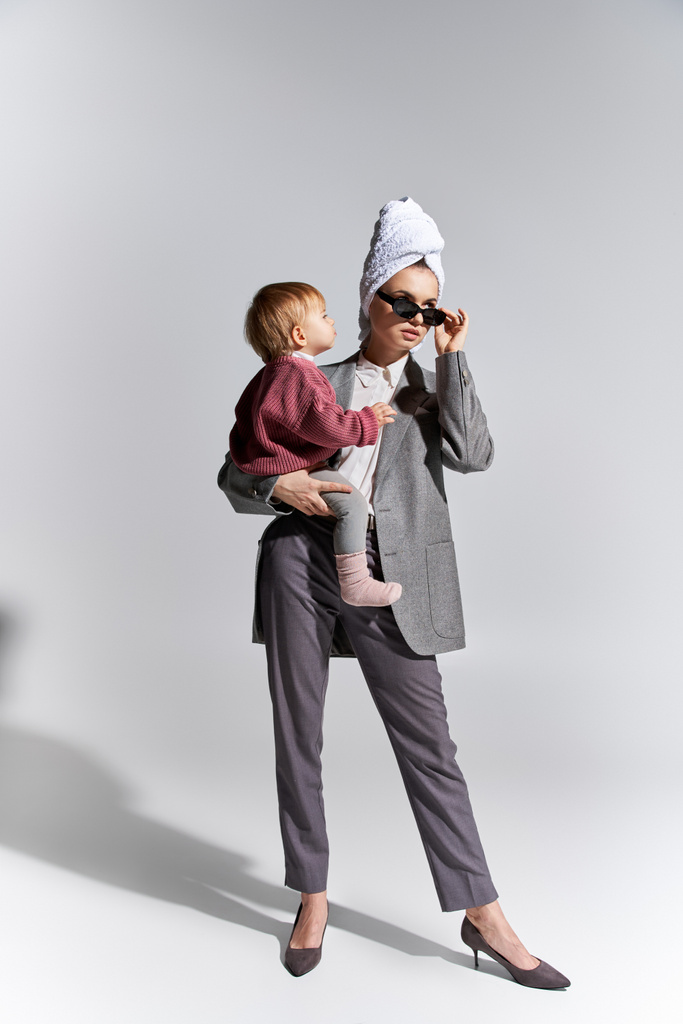 Frau mit Sonnenbrille, die Kleinkind-Tochter im Arm hält und mit Handtuch auf dem Kopf steht, Multitasking, ausgleichender Lebensstil, ermächtigte Frau in formeller Kleidung auf grauem Hintergrund, volle Länge  - Foto, Bild