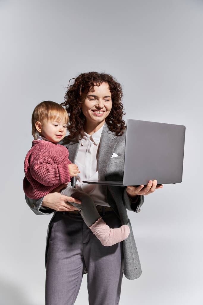 διαχείριση του χρόνου, ευτυχισμένη εργαζόμενη μητέρα που κρατά φορητό υπολογιστή και κόρη νήπιο στην αγκαλιά σε γκρι φόντο, την επαγγελματική ζωή αρμονία, την καριέρα και την οικογένεια, σύγχρονη ανατροφή των παιδιών, επαγγελματική επιτυχία, επιχειρηματίας  - Φωτογραφία, εικόνα