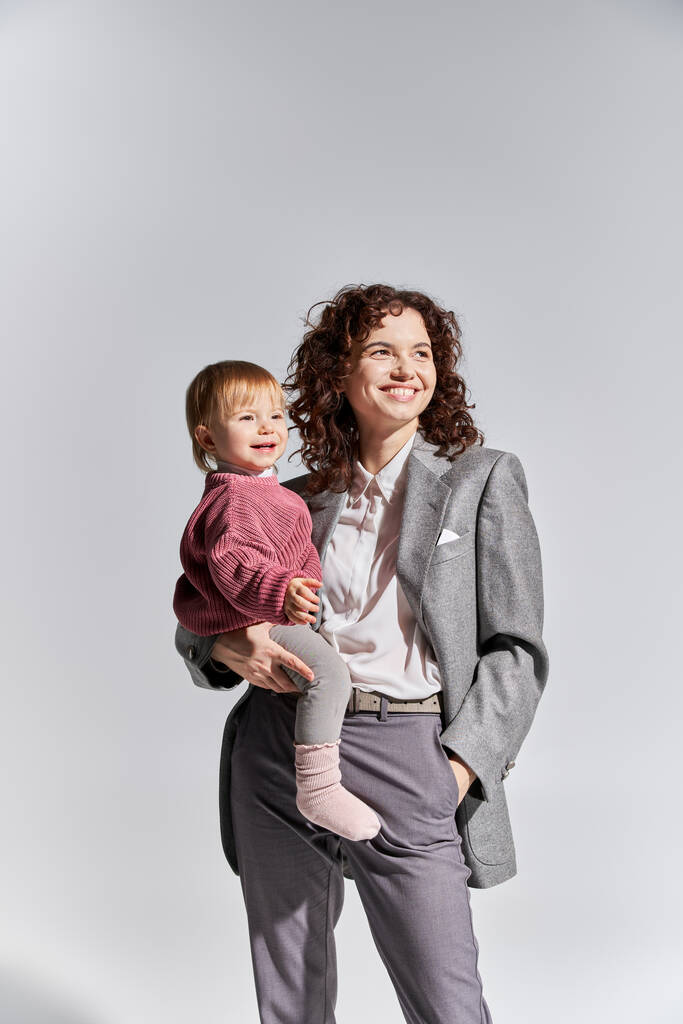 career and family, happy working mother holding toddler daughter in arms and standing with hand in pocket on grey background, work life balance, modern parenting, professional success, businesswoman  - Photo, Image